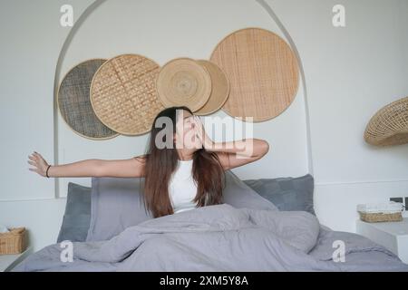 Giovane donna asiatica assonnata che sbadiglia e alza la mano svegliandosi la mattina presto dopo un sonno profondo, concetto di vacanza tranquilla al mattino Foto Stock