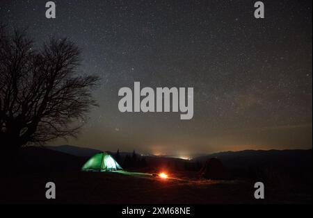 Campeggio notturno. La tenda solitaria si illumina dolcemente accanto al fuoco sotto il cielo stellato. La silhouette di un albero senza foglie si erge contro l'orizzonte montuoso, creando una serena e affascinante scena notturna in natura. Foto Stock