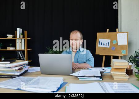 Un uomo con inclusività siede a una scrivania in un ufficio disordinato, concentrandosi sul suo notebook. Foto Stock