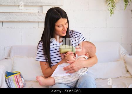 Madre premurosa che dà da mangiare al bambino con un biberon sul divano Foto Stock