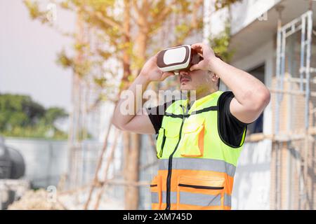 Un operaio edile che indossa un giubbotto di sicurezza utilizza un visore VR in un cantiere, illustrando la tecnologia nel settore edile. Foto Stock