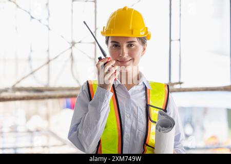 Lavoratrice edile che indossa indumenti di sicurezza e elmetto, comunica tramite radio e tiene i progetti in cantiere. Foto Stock