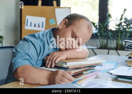 Un uomo con inclusività fa un pisolino alla sua scrivania, appoggiando la testa su una pila di documenti. Foto Stock