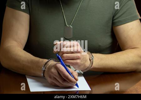 Un militare siede ad una scrivania, ammanettato, firma un documento su carta bianca in una stanza poco illuminata. Foto Stock