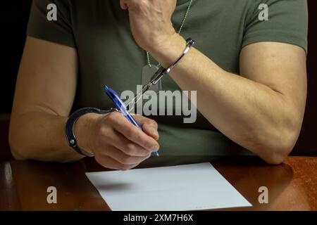 Un militare siede ad una scrivania, ammanettato, firma un documento su carta bianca in una stanza poco illuminata. Foto Stock