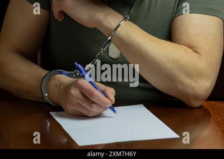 Un militare siede ad una scrivania, ammanettato, firma un documento su carta bianca in una stanza poco illuminata. Foto Stock