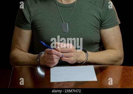 Un militare siede ad una scrivania, ammanettato, firma un documento su carta bianca in una stanza poco illuminata. Foto Stock