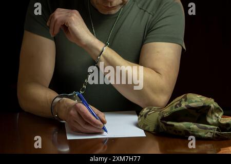 Un militare siede ad una scrivania, ammanettato, firma un documento su carta bianca in una stanza poco illuminata. Foto Stock