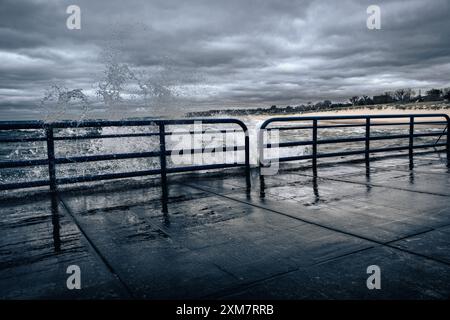 Una potente onda oceanica si schianta contro una robusta ringhiera posizionata lungo il bordo dell'acqua. Foto Stock