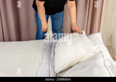 Una donna dispone meticolosamente una coperta bianca su un letto, soffici cuscini per creare un ambiente accogliente. Il letto non fatto presenta lenzuola leggermente spiegazzate, una Foto Stock