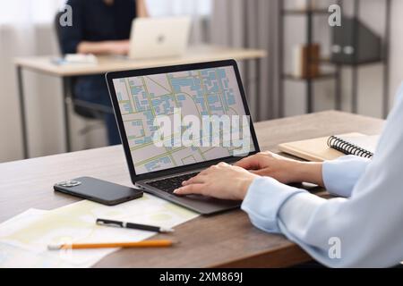 Cartografo che lavora con una mappa catastale su un computer portatile al tavolo di legno in ufficio, primo piano Foto Stock