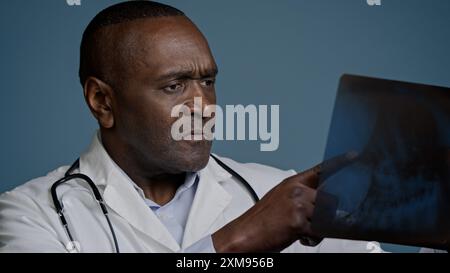 Medico maschio afroamericano pensivo maturo esaminare radiografia in sfondo blu studio uomo chirurgo traumatologo controllare risultati scansione analizzare lesioni su Foto Stock