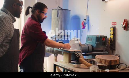 Produttore che insegna ai nuovi dipendenti come sagomare la ciotola di legno sulla levigatrice a disco nell'officina di assemblaggio. Apprendista che impara da uno specialista nella lavorazione del legno come utilizzare il disco rotante della macchina per la molatura, telecamera B Foto Stock