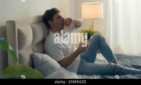Uomo caucasico sdraiato su letto casa da letto confortevole accogliente sbadiglio pigrizia maschio annoiato assonnato tenendo cellulare smartphone SMS chat Internet Foto Stock