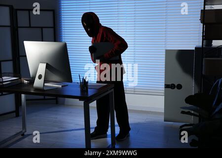 Ladro che indossa balaclava per rubare un laptop in ufficio di notte. Rapina Foto Stock