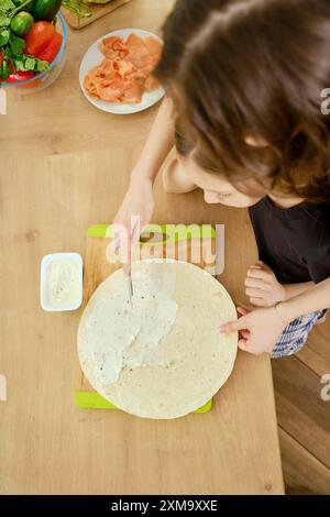 Vista dall'alto di una donna che spalma formaggio spalmabile su una focaccia mentre la figlia la guarda e impara a cucinare Foto Stock