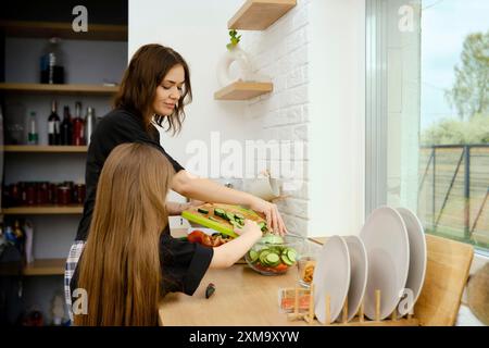 La bambina aiuta la mamma a mettere il cetriolo tritato in una ciotola con insalata Foto Stock