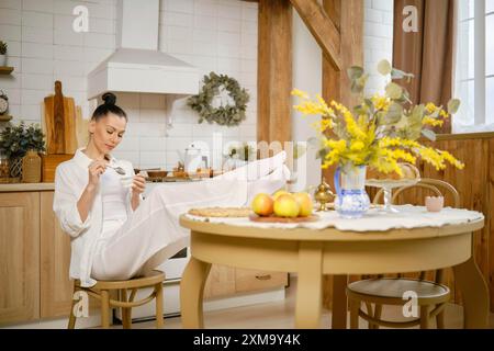 Una donna in abito di cotone si siede in cucina con i piedi sul tavolo e mangia yogurt Foto Stock