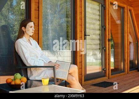 Donna serena in abito bianco che legge un libro su una moderna veranda, con succo d'arancia e frutta fresca nelle vicinanze Foto Stock