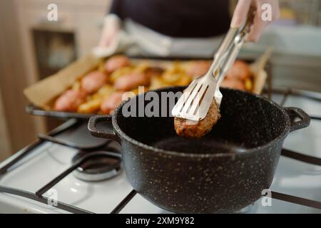 La vista ravvicinata di una donna irriconoscibile mette la cotoletta di carne in una pentola usando le pinze da cucina Foto Stock