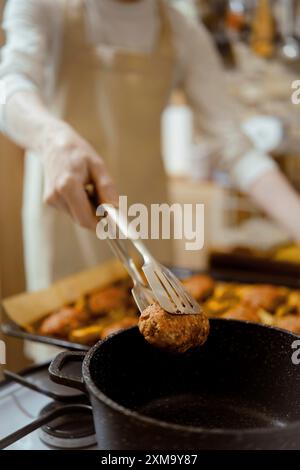 Vista ravvicinata del trasferimento delle polpettine di carne da un vassoio da forno a una pentola con pinze da cucina Foto Stock