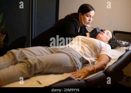 Un professionista di bellezza che si concentra sul viso di una donna durante un trattamento mentre si sdraia, procedura di tatoo permanente delle sopracciglia microblante, beauty spa Foto Stock