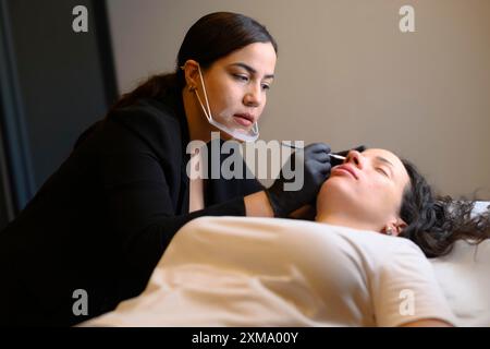 Cosmetologo in guanti neri e una maschera che esegue un trattamento sopracciglia con una matita su una donna sdraiata, microblading sopracciglia permanente Foto Stock