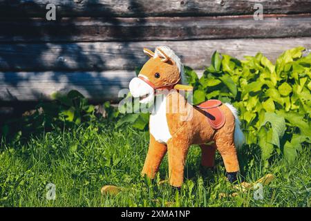 Un cavallo giocattolo imbottito si erge su un'erba verde di fronte a una recinzione di legno e la luce del sole splende su di essa Foto Stock