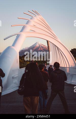 Persone che attraversano un ponte artistico al tramonto, con una montagna innevata sullo sfondo, il Monte Taranaki, nuova Zelanda Foto Stock