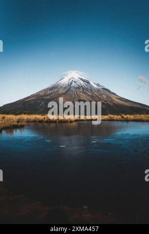 Una montagna innevata che si riflette in un lago fermo sotto un cielo azzurro, il Monte Taranaki, nuova Zelanda Foto Stock