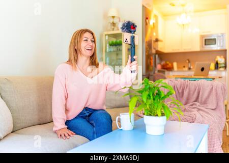 Donna bionda adulta, vlogger in streaming da casa utilizzando un telefono cellulare seduto in salotto Foto Stock