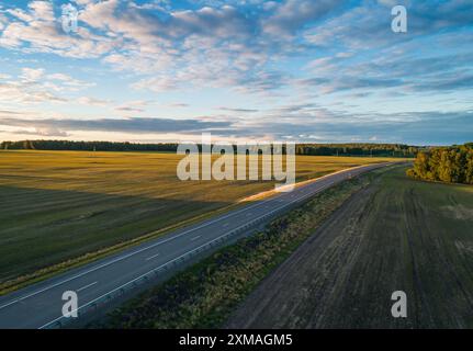 Una strada tortuosa si estende in lontananza, fiancheggiata da vivaci campi verdi e fitti alberi sotto un cielo azzurro. Foto Stock