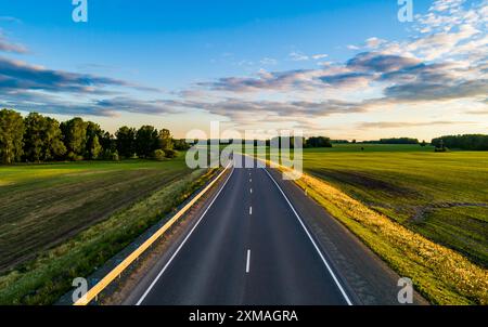 Una strada tortuosa si estende in lontananza, fiancheggiata da vivaci campi verdi e fitti alberi sotto un cielo azzurro. Foto Stock