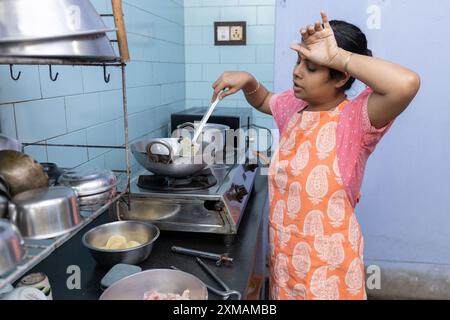 Una casalinga indiana che indossa un grembiule da cucina per preparare il cibo e stancarsi con gli utensili in alluminio su un forno a gas Foto Stock