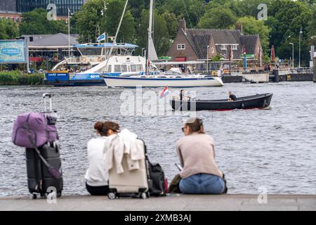 Turisti seduti sul retro della stazione ferroviaria centrale di Amsterdam, sulle rive del fiume IJ, Amsterdam, Paesi Bassi Foto Stock