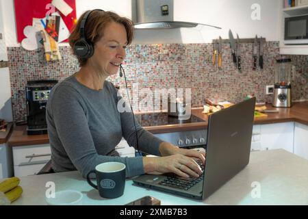 Donna, metà degli anni '50, lavora da casa, con un computer portatile e comunica con i colleghi tramite cuffie, ufficio domestico, cucina Foto Stock