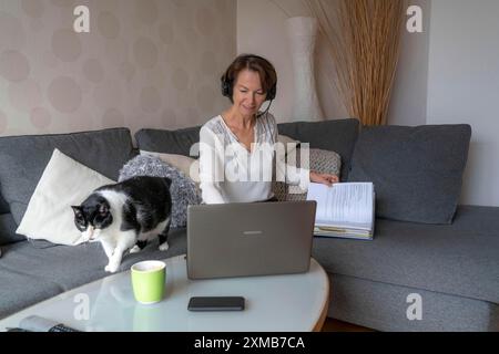 Donna, metà degli anni '50, lavora da casa, con un computer portatile e comunica con i colleghi tramite cuffie, ufficio domestico o divano Foto Stock