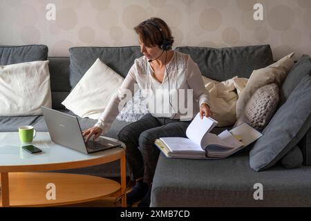 Donna, metà degli anni '50, lavora da casa, con un computer portatile e comunica con i colleghi tramite cuffie, ufficio domestico o divano Foto Stock