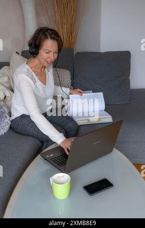 Donna, metà degli anni '50, lavora da casa, con un computer portatile e comunica con i colleghi tramite cuffie, ufficio domestico o divano Foto Stock
