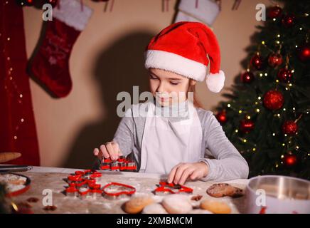 Una ragazza con un cappello di Babbo Natale lavora con dei taglierini per tagliare i biscotti di Natale. Foto Stock