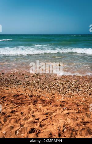 Vivace scenario di spiaggia con sabbia arancione, ciottoli e onde blu dell'oceano sotto un cielo limpido. Perfetto per vacanze estive. Foto Stock