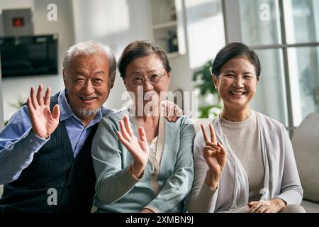 gruppo di tre persone asiatiche anziane felici sedute sul divano di casa che saltano le mani alla macchina fotografica mentre chiacchierano video Foto Stock