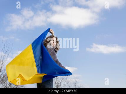 Giovane donna, una ragazza adulta, ha una grande bandiera Ucraina giallo-blu dietro la schiena contro il cielo. Sostieni l'Ucraina. Sono orgoglioso di essere ucraino. Inde Foto Stock