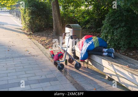 Un senzatetto dorme su una panchina del parco con il suo camminatore davanti a lui Foto Stock