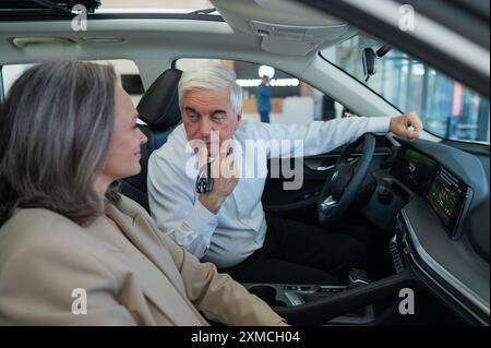 Un uomo anziano tiene le chiavi mentre è seduto in una macchina nuova. Una coppia sposata dai capelli grigi è felice di comprare un'auto. Foto Stock
