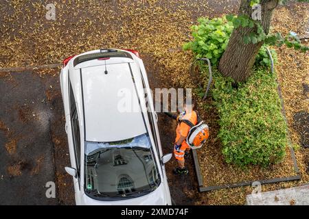 Pulizia stradale, dipendente del servizio municipale di pulizia della città, pulisce marciapiedi e parcheggi, con un soffiatore di foglie, in estate, dagli alberi Foto Stock