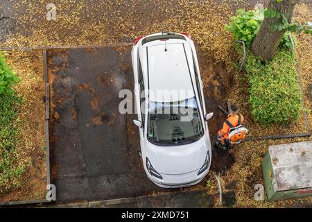 Pulizia stradale, dipendente del servizio municipale di pulizia della città, pulisce marciapiedi e parcheggi, con un soffiatore di foglie, in estate, dagli alberi Foto Stock