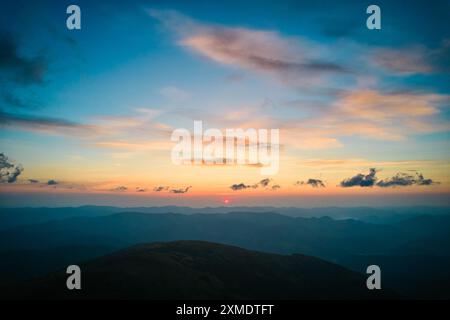 Il sole tramonta dietro le colline ondulate, gettando un caldo bagliore dorato nel cielo. Le nuvole mostruose, tinto da sfumature di arancio e giallo, si estendono su un cielo blu profondo. Paesaggio sereno ed esteso. Foto Stock