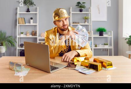 Ritratto dell'uomo in tuta dorata che tiene in mano pile di contanti e lavora su un notebook Foto Stock