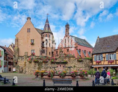 La fontana di St. Leon in Piazza Saint-Leon nel centro di Eguisheim in Alsazia. Case ben tenute e in legno e decorazioni floreali caratterizzano Foto Stock